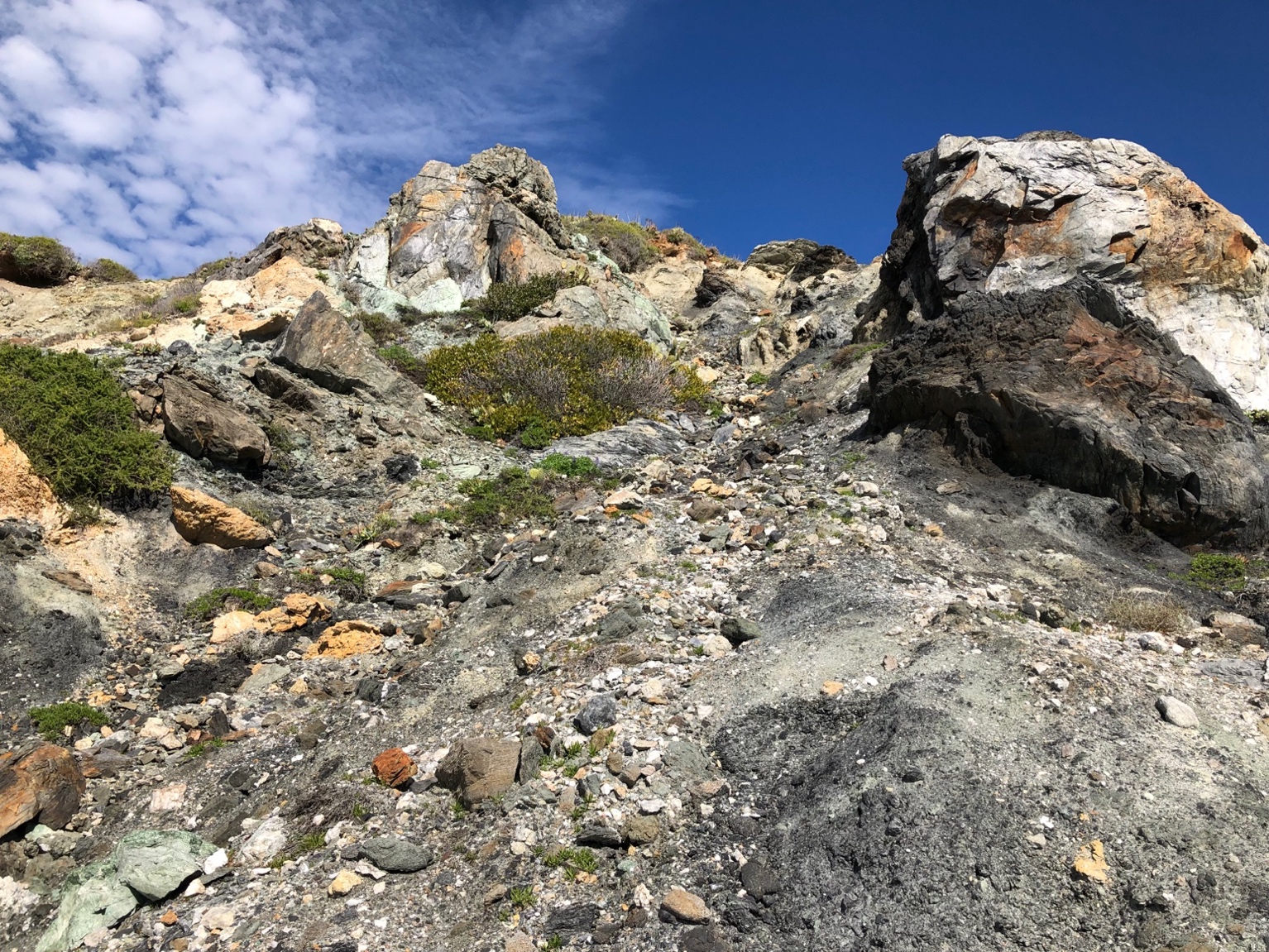 A rocky hill with blue sky and clouds. he rubble hill is right behind the whale tail has a lot of interesting rocks on it. These were transported down the streams forming a talus slope of mixed rocks.Description automatically generated